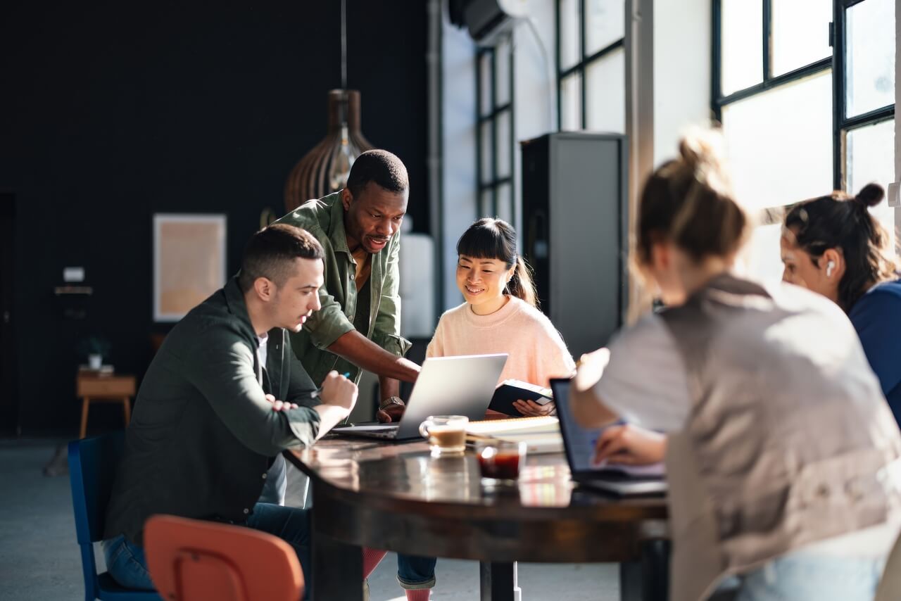 Group of five people collaborating around a table with laptops and coffee in a bright workspace.