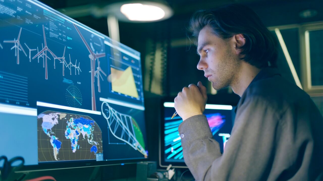 Person analyzing data on multiple monitors showing wind turbines, global maps, and technical graphs.